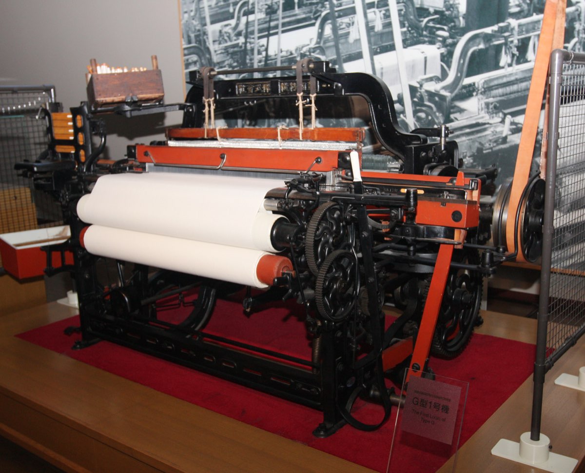 A black iron loom with orange-red parts and rolls of white cloth, on a red base in a museum display, with a label in front reading "G型1号機 The First Loom of Type G" and an old photograph of a weaving shed on the wall behind