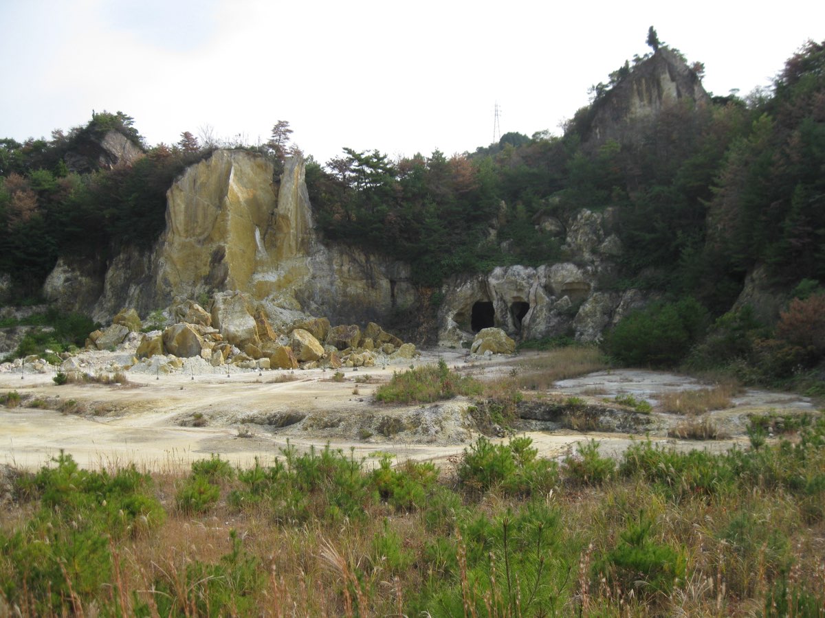 The Izumiyama quarry in Arita - a pale, flat quarry floor with grass in the foreground, a tall cut face of yellowish rock with fallen boulders at its foot, two dark tunnel openings in the rock on the right, and wooded hills behind