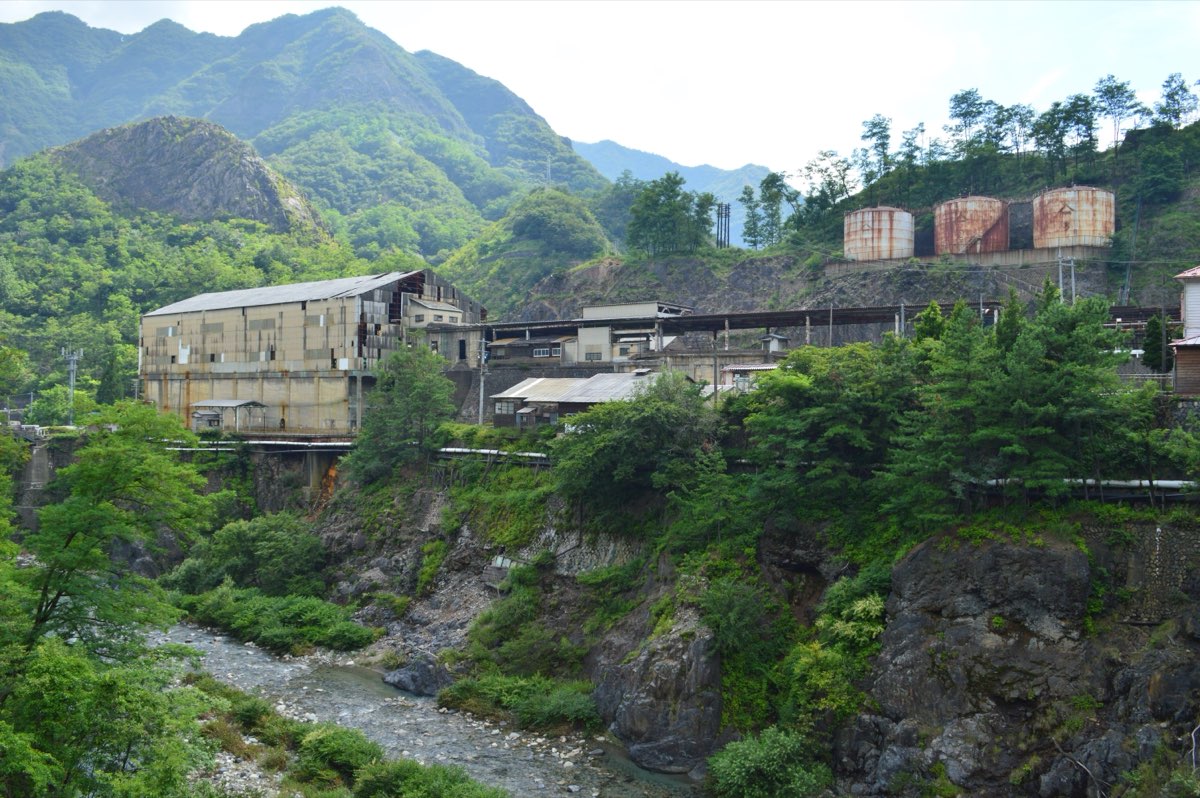 Weathered factory buildings and three rusted round tanks of the former Ashio copper mine smelter on a slope above a rocky river, with green forested mountains and a bare rocky peak behind