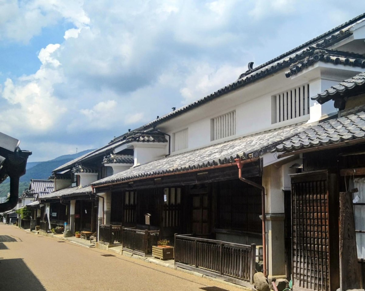 A quiet street of two-storey townhouses in Wakimachi with white plastered upper walls, dark wooden lattices, grey tiled roofs and protruding firewalls topped with tiles, under a cloudy blue sky with green hills behind