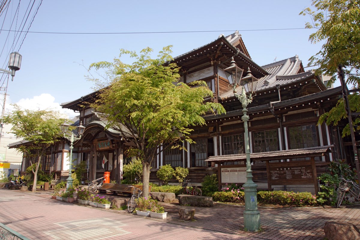 The two-storey wooden building of Takegawara Onsen in Beppu, with tiled roofs and a curved gable over the entrance, seen from a paved street with young trees, street lamps, flower planters and parked bicycles under a blue sky