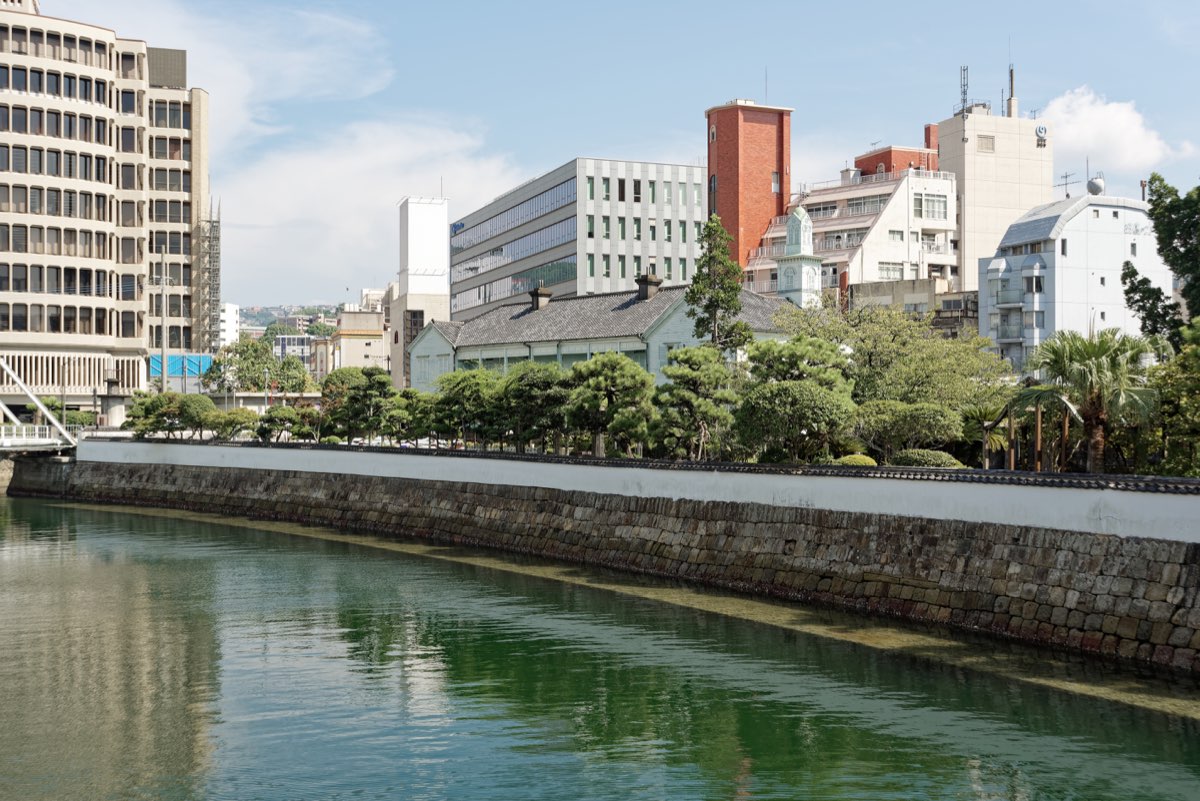 A stone embankment topped by a long white wall runs along a green river; behind it are pine trees and a white building with a grey tiled roof, with modern city buildings rising in the background under a blue sky