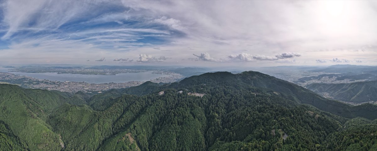 Wide aerial view over the forested ridges of Mount Hiei, with a few buildings among the trees, Lake Biwa and the city of Ōtsu spreading along its shore on the left, under a cloudy sky