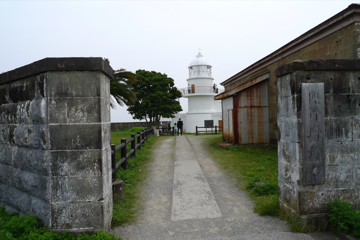 A white lighthouse seen at the end of a paved path between old stone gateposts, with a low stone building on the right, a tree on the left and a hazy sky