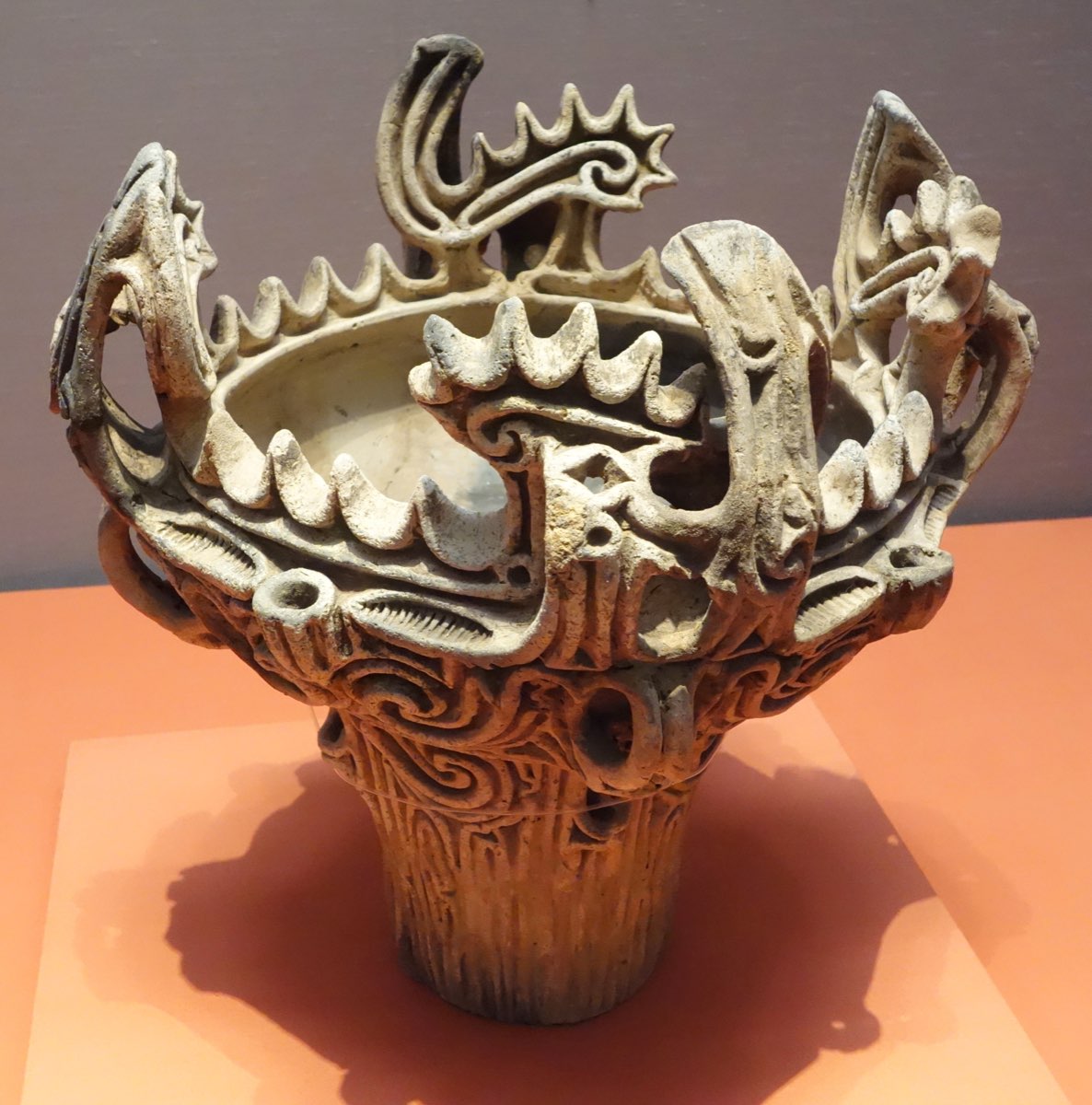 A flame-style clay vessel on an orange museum plinth, its rim crowded with tall curling projections and rows of sawtooth points, and its body covered with swirling raised lines
