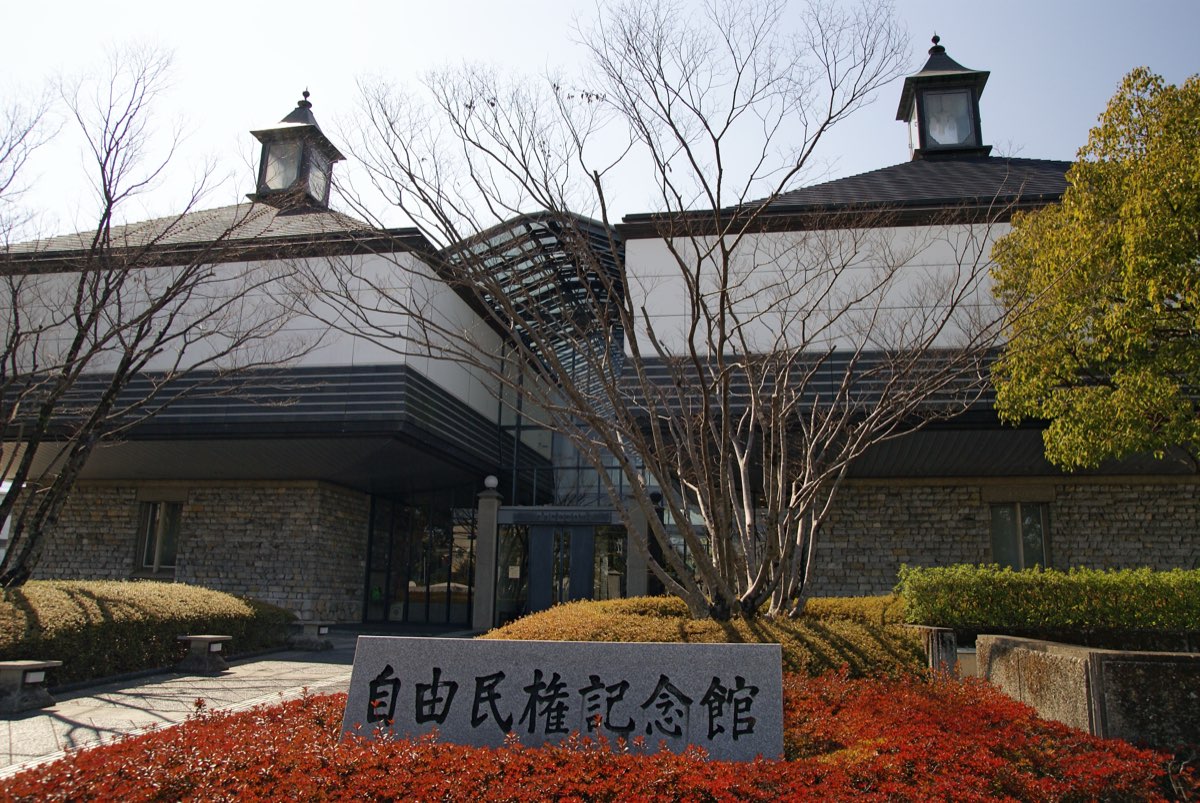 Front of the Kōchi City Freedom and People's Rights Museum, a building with stone-faced lower walls, white upper walls and two lantern-like roof turrets, behind bare trees, hedges and a stone sign reading 自由民権記念館 among red shrubs