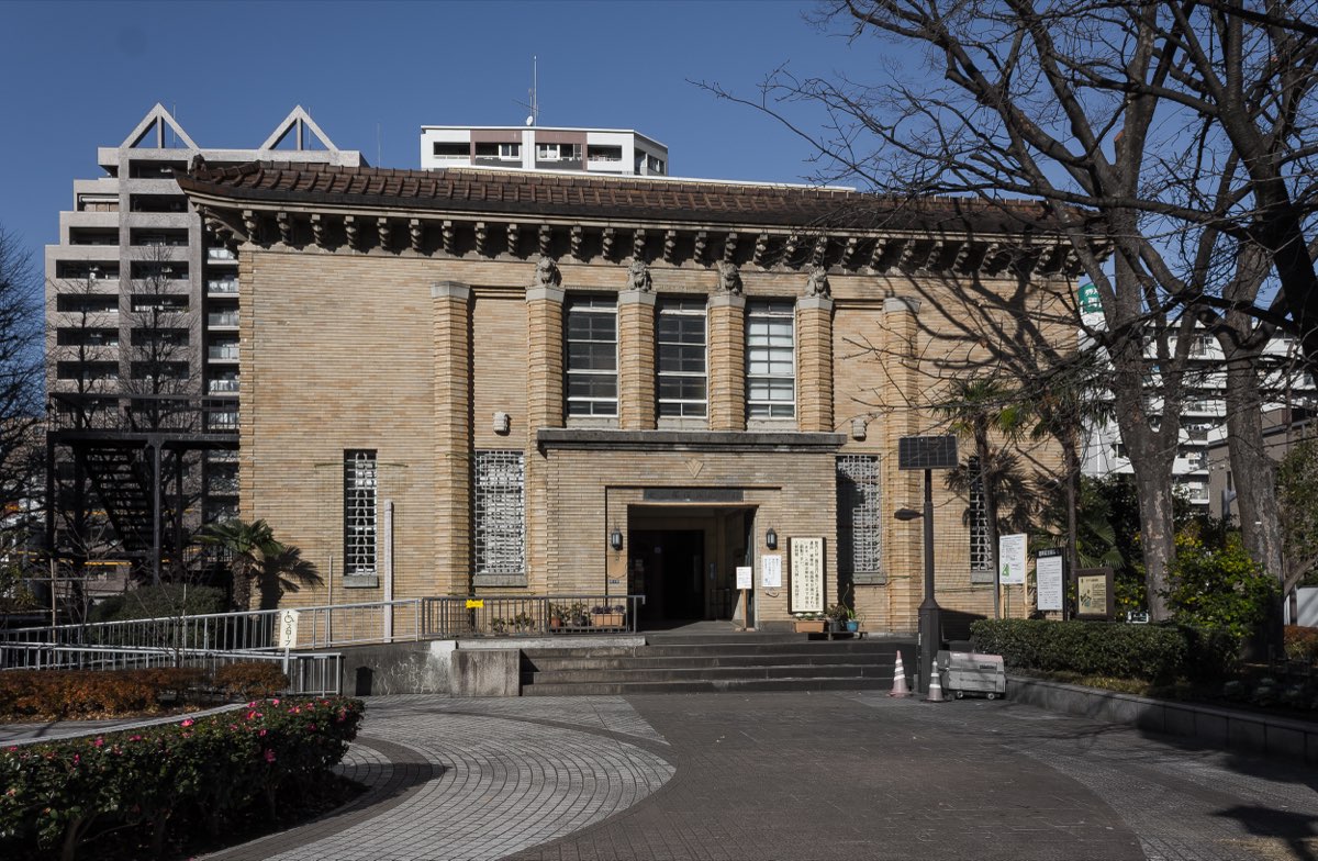 A building faced with brown tiles, with a tiled roof, tall pilasters topped by small sculpted figures, and a central entrance up a short flight of steps, under a clear blue sky in a park