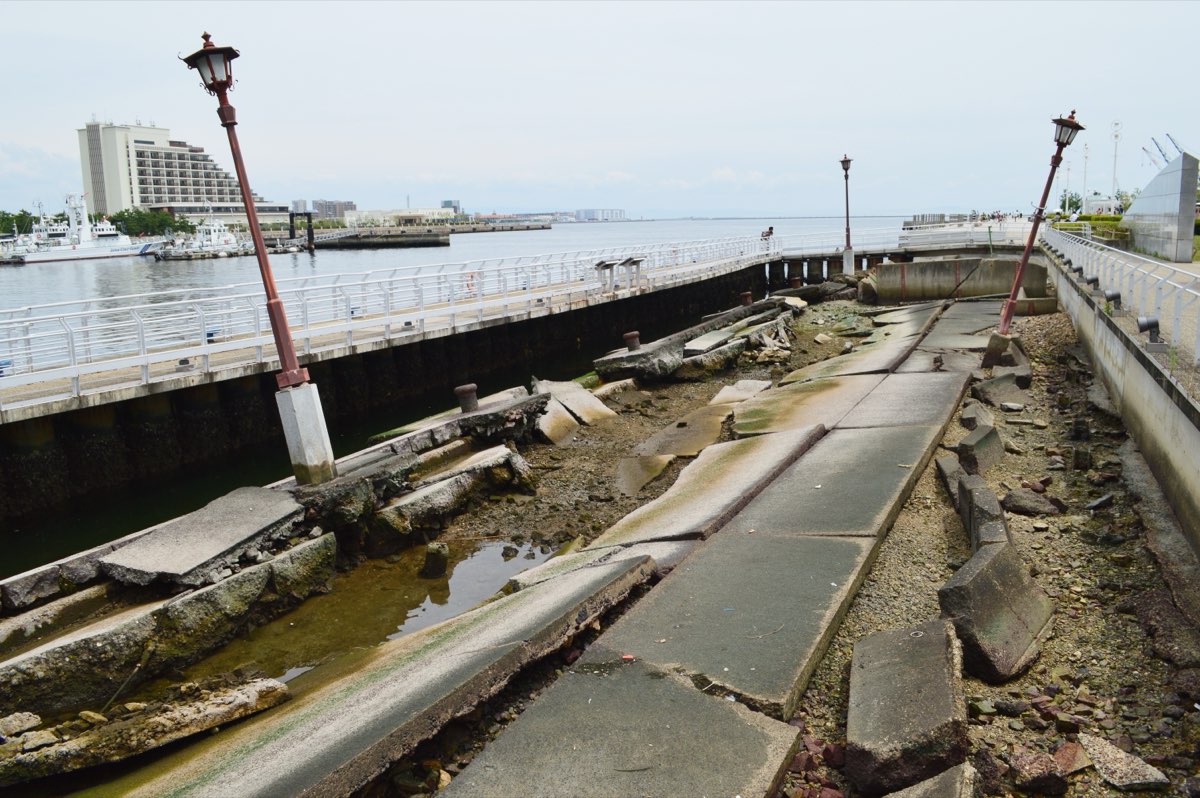 A broken concrete quay with tilted and cracked slabs and fallen blocks, two old street lamps leaning at an angle, a walkway with white railings beside it, and the harbour with a hotel and boats in the background under a pale sky