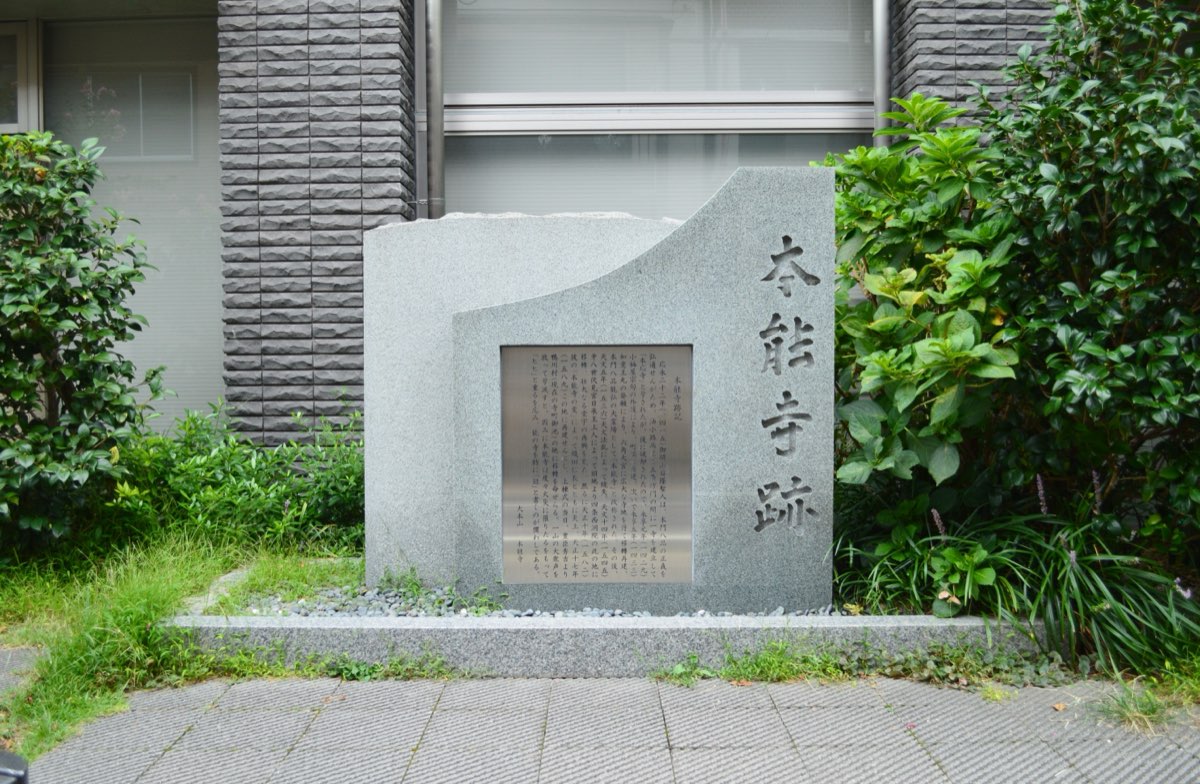 A grey granite monument on a paved walkway in front of a modern building, with the characters for "Honnō-ji site" carved vertically on the right and a metal plaque of Japanese text set into the stone, framed by green shrubs