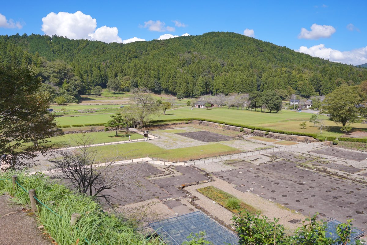 View from a slope over the excavated site of the Asakura residence at Ichijōdani, with gravel-covered building plots, rows of foundation stones, low stone walls and lawns, and a forested hill behind under a blue sky