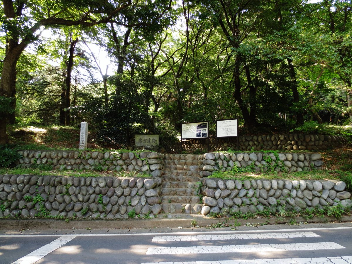 Point A of the Iwajuku site in 2016, a wooded slope faced with low terraces of rounded stones and central steps, with a stone pillar, a stone plaque and two information boards, seen across a road with a pedestrian crossing