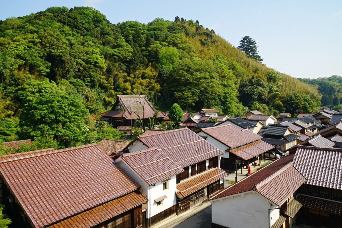 View over the tiled roofs of traditional two-storey wooden houses along a narrow street in Ōmori, with a larger tiled roof and a steep forested hillside behind under a clear sky