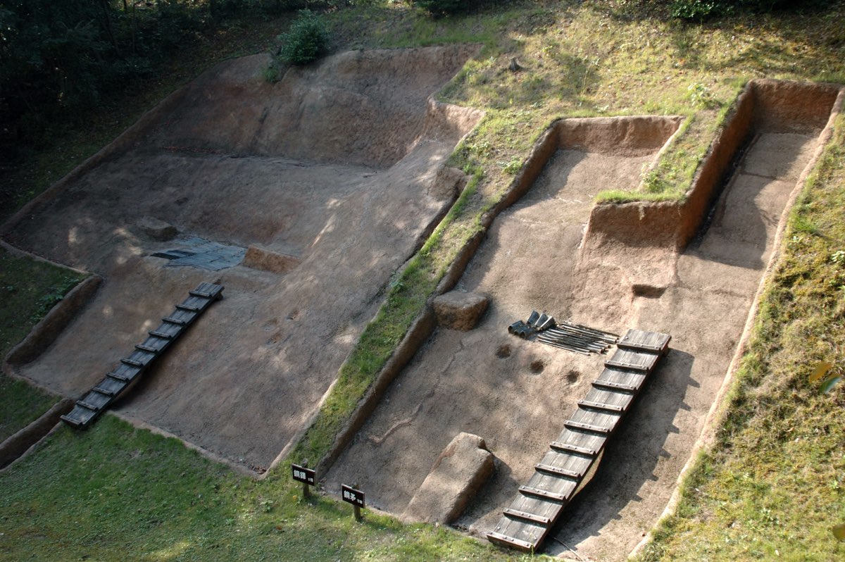 Two stepped excavation trenches cut into a grassy hillside, with low wooden walkways and, in one trench, a group of dark objects laid on the bare earth to show where bronzes were found
