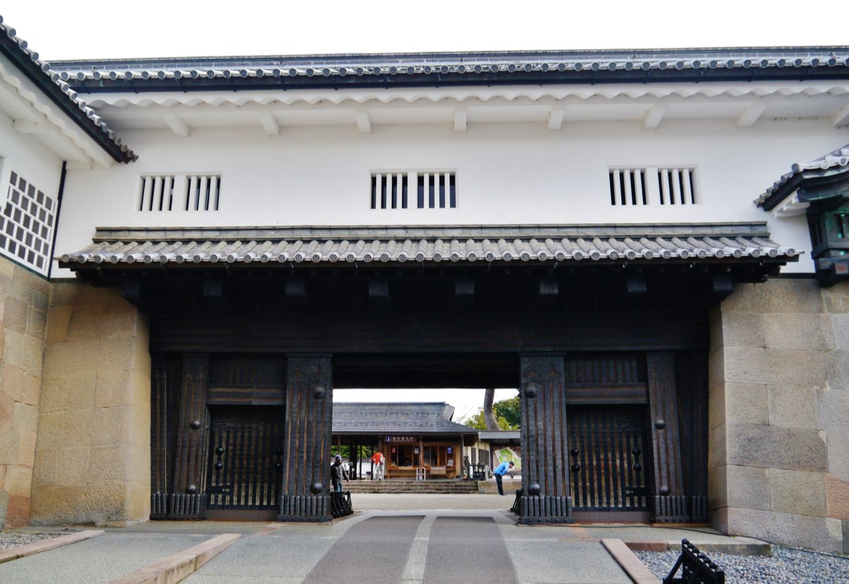 The Ishikawa Gate of Kanazawa Castle seen from the front - a large dark wooden gate set between walls of fitted stone blocks, under a white-plastered upper storey with barred windows and grey tiled roofs; a paved path leads through the open gate to a courtyard with a small building beyond