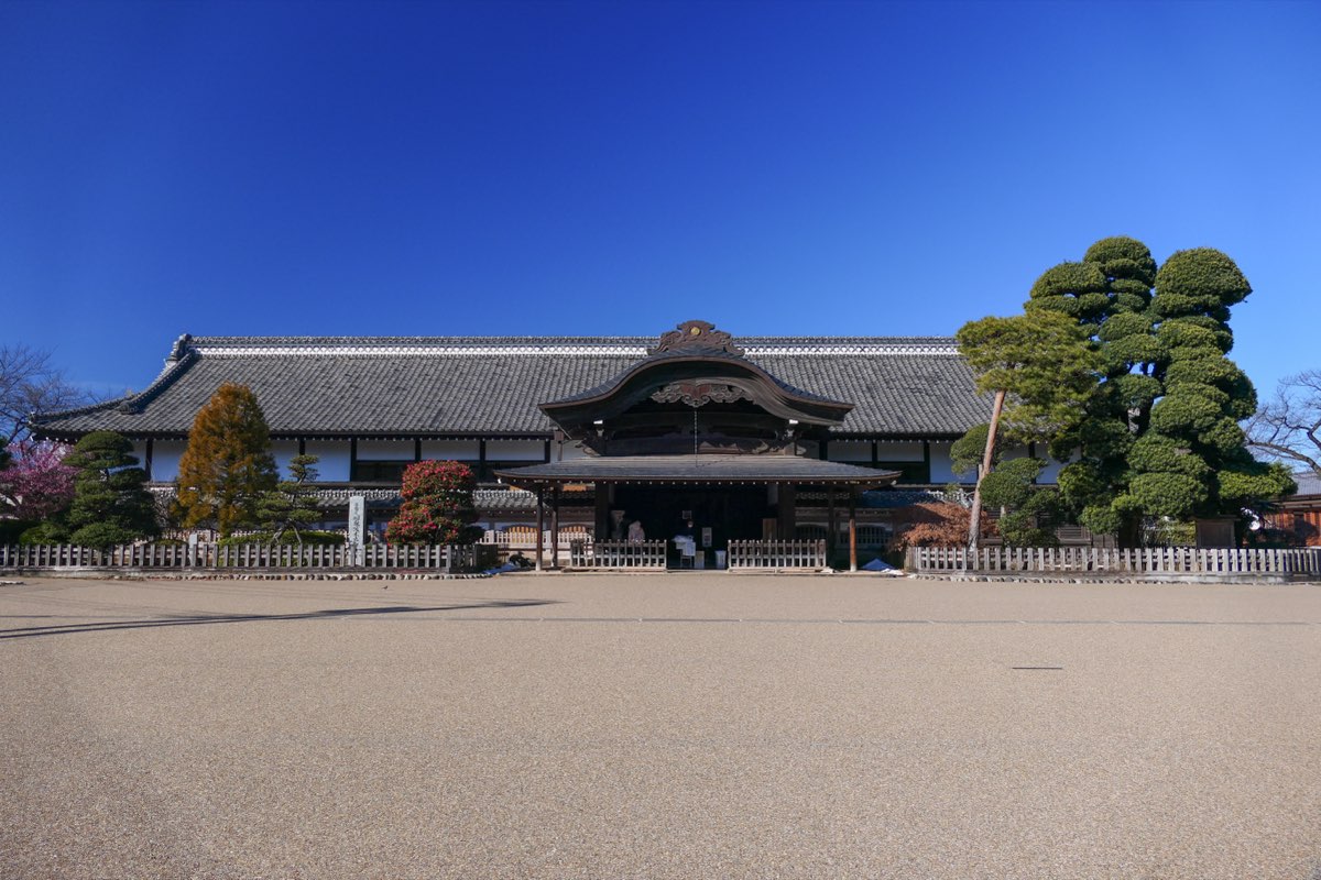 The front of the Honmaru Goten of Kawagoe Castle, a long single-story building with a grey tiled roof and a curved gable over the entrance, behind a low wooden fence and a wide gravel forecourt under a clear blue sky, with pruned pines and other trees at the sides