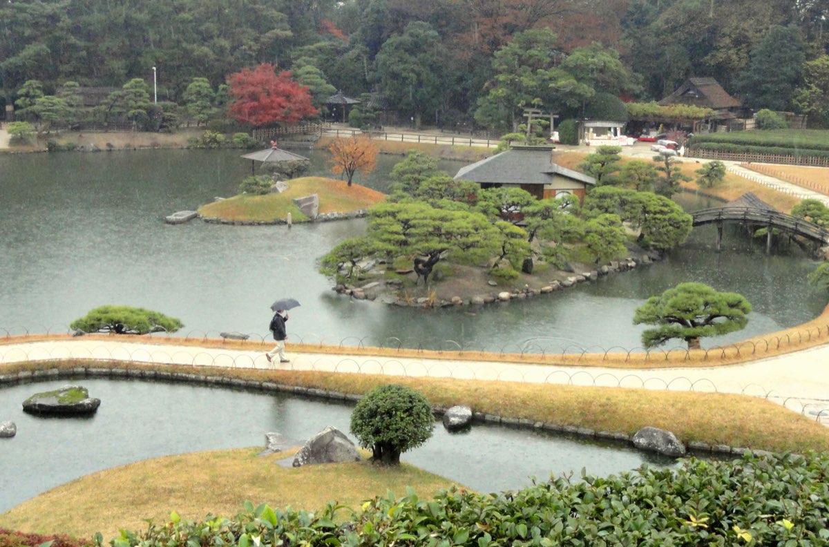 A wide pond in Kōrakuen seen from a height on a rainy day, with a pine-covered island holding a small roofed building, a second grassy island with an orange-leaved tree and an umbrella-shaped shelter, a red-leaved tree on the far shore, an arched wooden bridge on the right, a path between two ponds in front where one person walks with an umbrella, and wooded hills behind