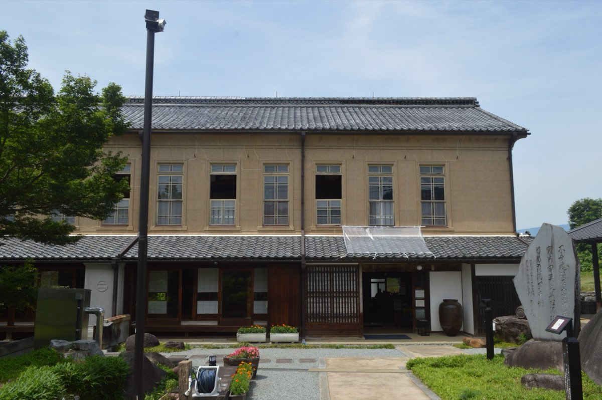A two-storey building with a tiled roof, a beige upper floor with rows of tall windows and a traditional wooden ground floor, with a large inscribed stone and trees in front under a blue sky