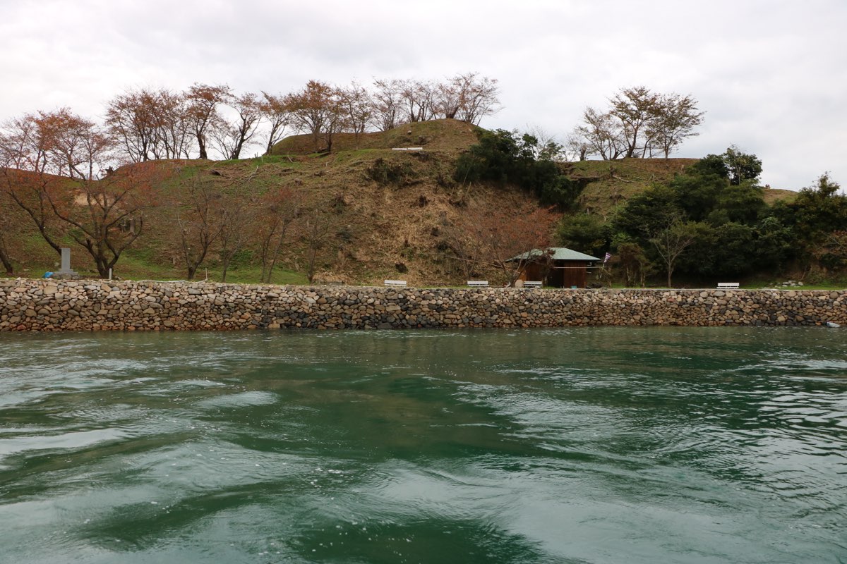 The island of Noshima seen from a boat, with swirling green water in front, a long stone wall along the shore, and grassy terraced slopes with bare trees rising to a flat top under a cloudy sky