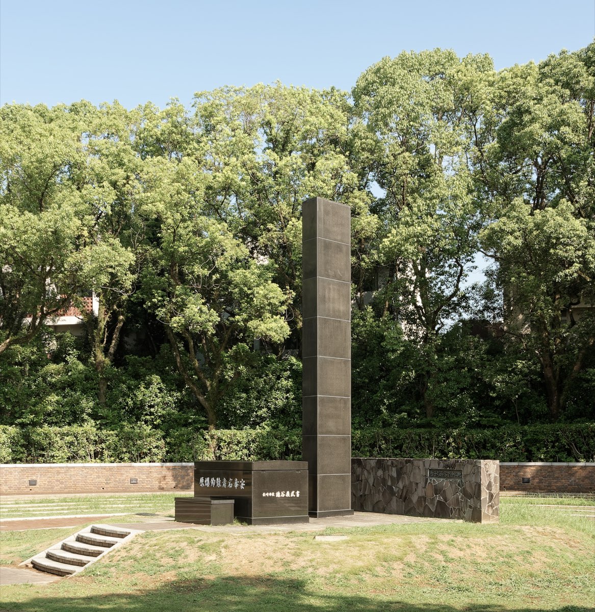 A tall, slender dark stone pillar stands on a grassy mound in a park, with a low dark stone block inscribed in Japanese at its base, a low wall of irregular stones beside it, and leafy green trees behind under a clear sky