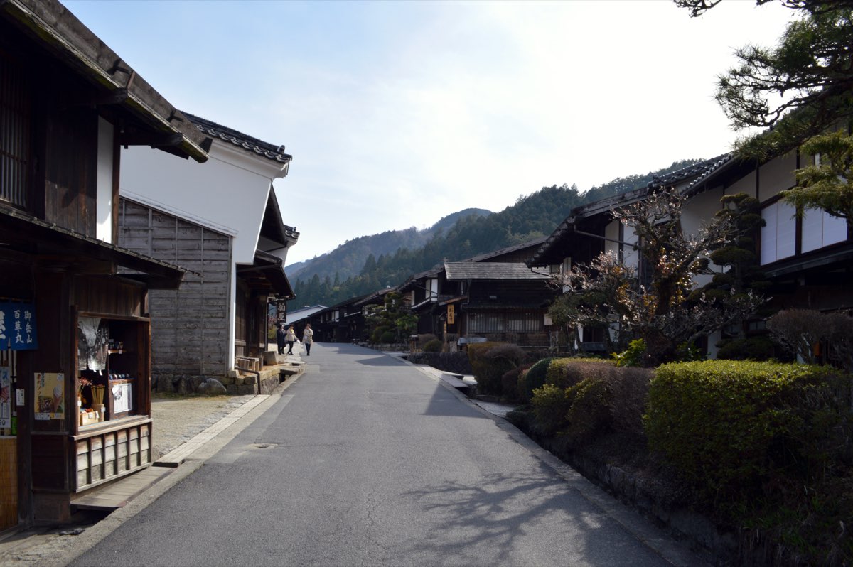 A quiet paved street lined with dark wooden two-storey houses and white plastered walls, with clipped shrubs on the right and forested hills behind, photographed in March 2016
