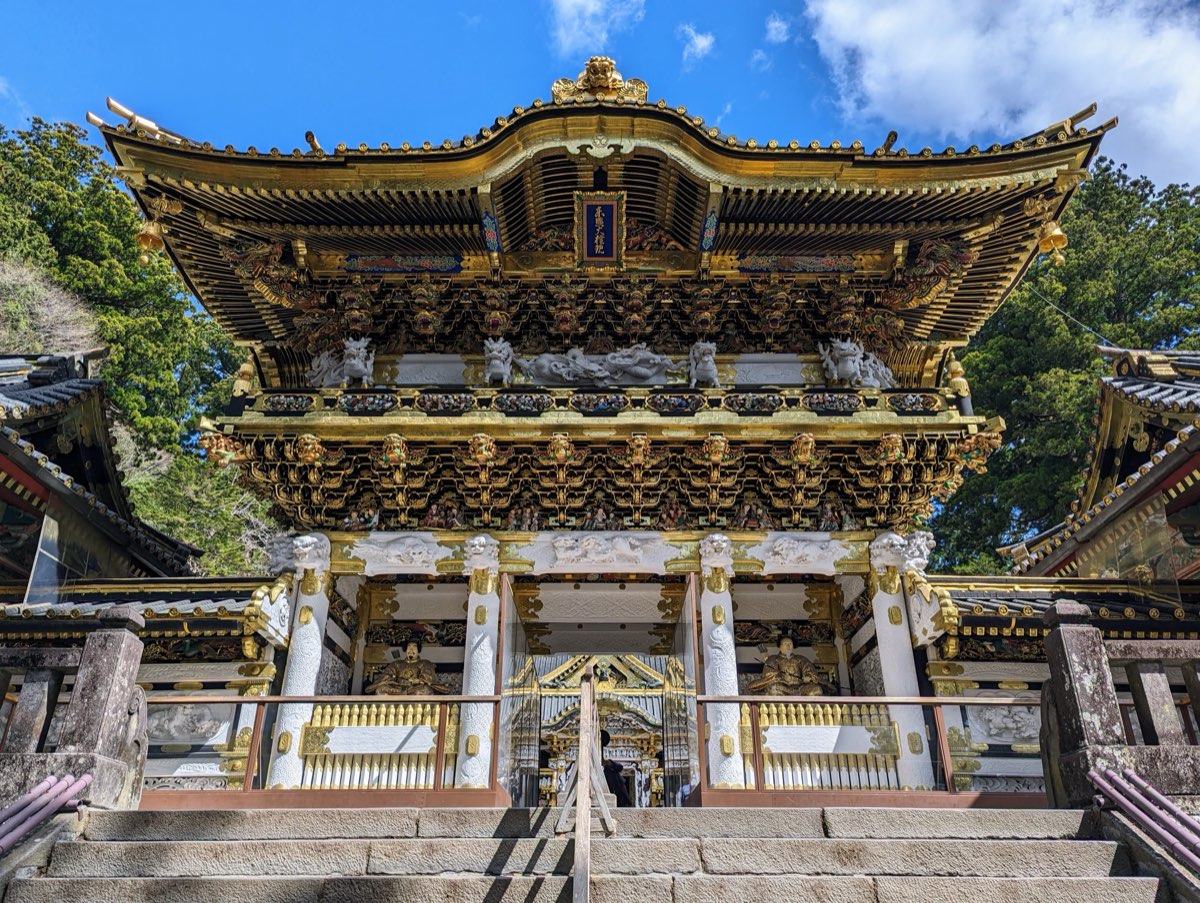 The Yomeimon gate of Nikko Toshogu seen from the foot of its stone steps, a two-storey gate with white columns, gilded brackets and dense colored carvings under a curved black-and-gold roof, with cedar trees and blue sky behind