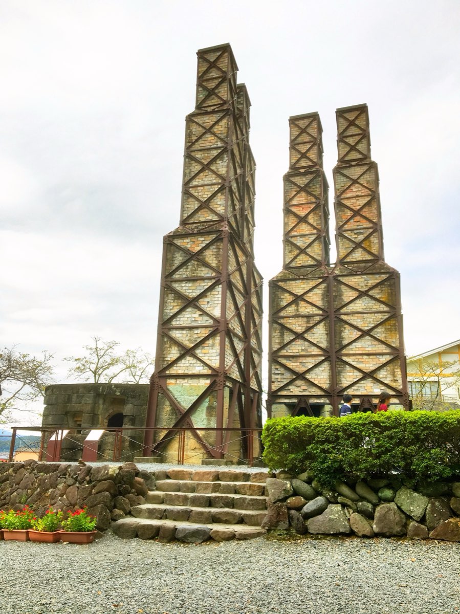 Two tall square brick chimney structures, each pair of chimneys braced by a criss-cross frame of reddish-brown steel trusses, rising from stone furnace bodies behind stone steps, a rough stone wall and a trimmed hedge, with a low stone structure on the left and a cloudy sky above; two people stand small at the foot of the right-hand structure