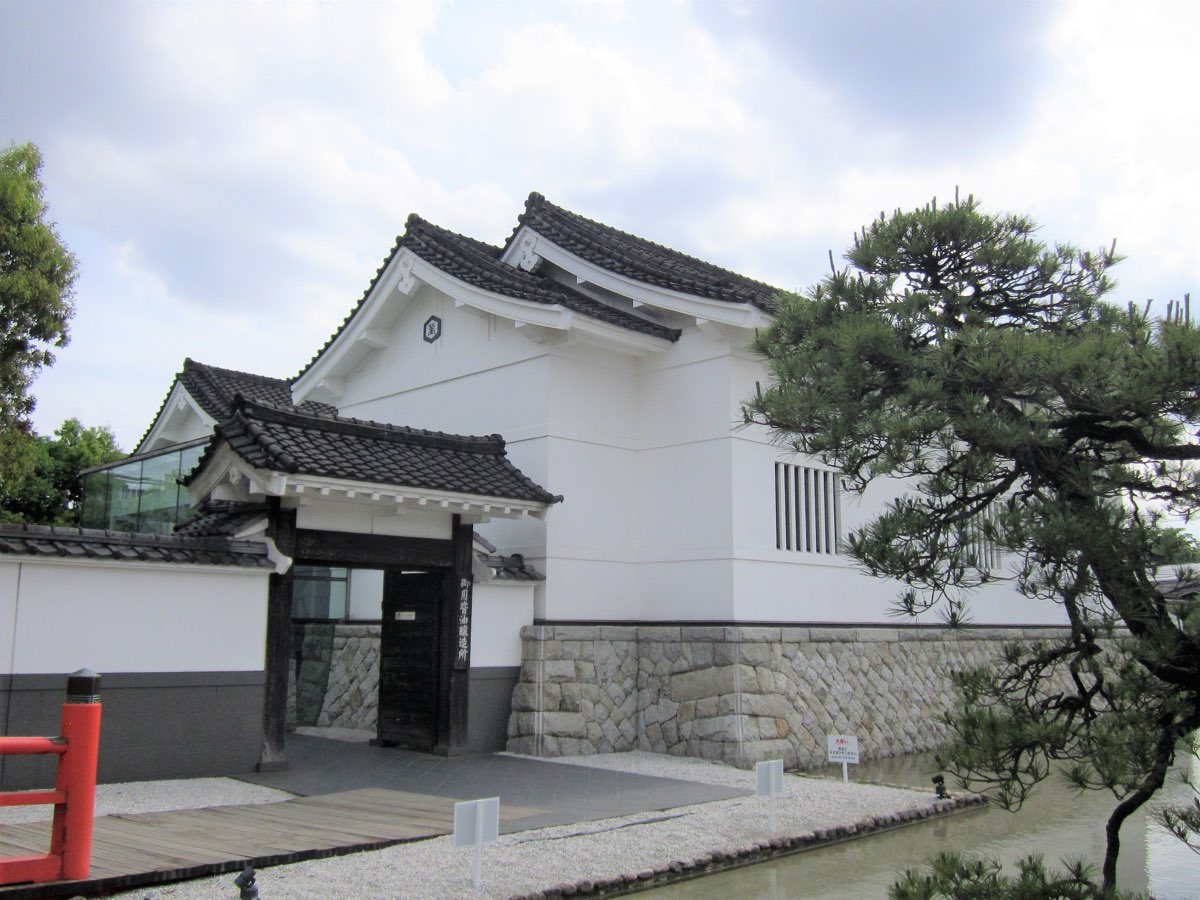 A white-walled storehouse with a tiled roof and a stone base behind a gate, with a pine tree at the front right, at the Kikkoman factory in Noda, photographed in May 2019