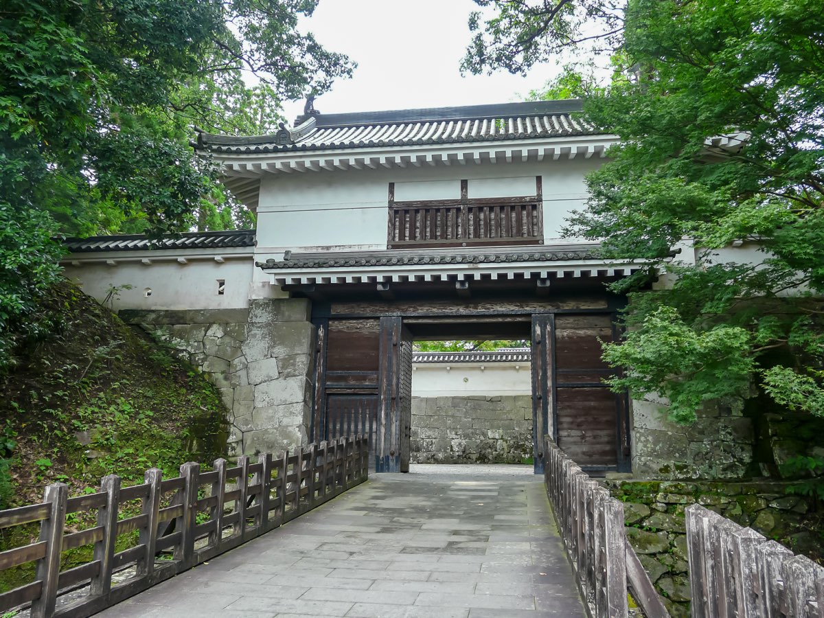A white-walled two-storey gatehouse with a tiled roof and heavy wooden doors standing open, set on mossy stone walls among green trees, reached by a paved path with wooden railings