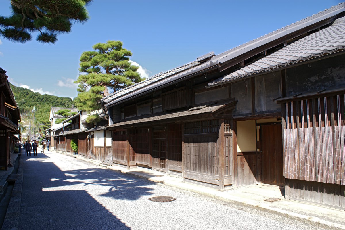 A quiet street of old two-storey wooden townhouses with dark lattice fronts and tiled roofs, a pine tree rising behind a wall, wooded hills in the distance and a blue sky
