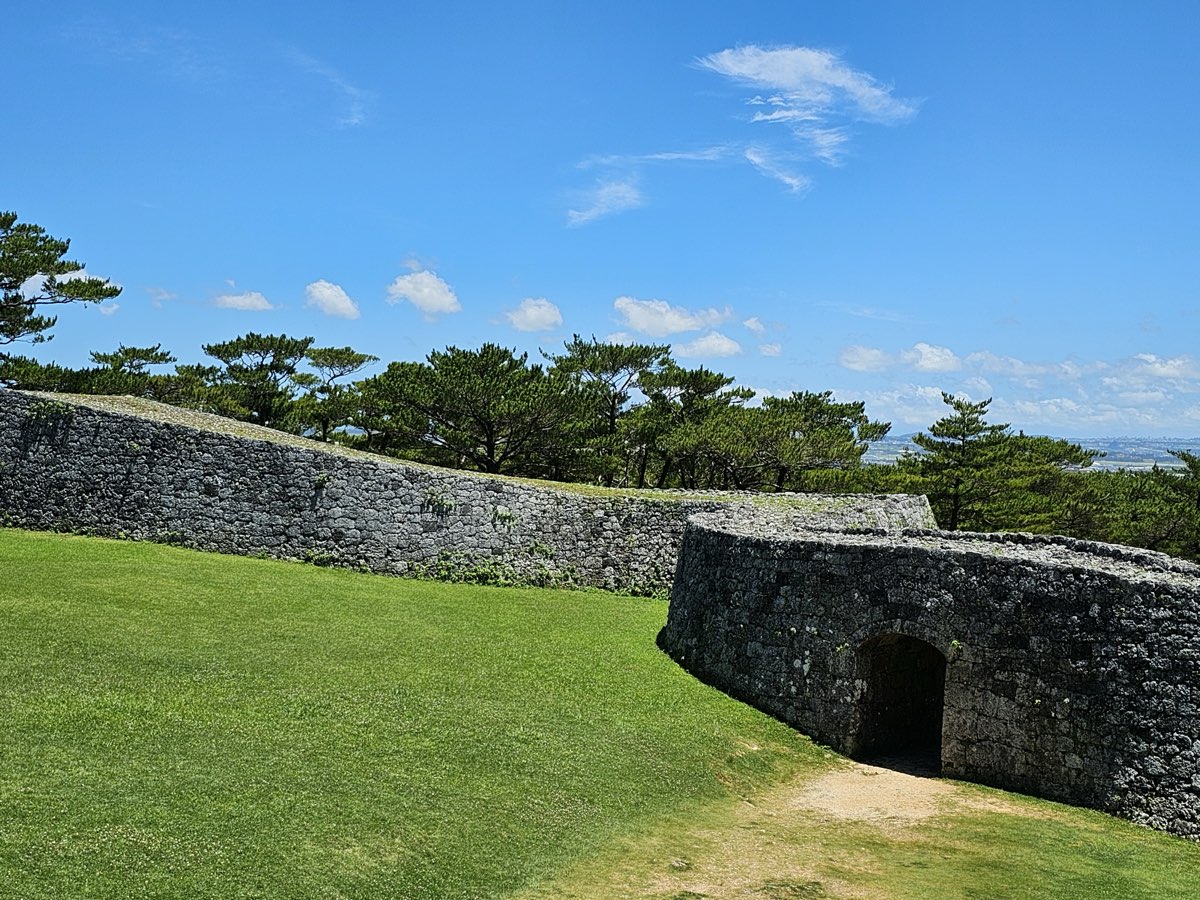 Curving grey stone walls of Zakimi Castle on a grassy hilltop, with an arched gateway in the wall on the right, pine trees behind and a blue sky with scattered clouds