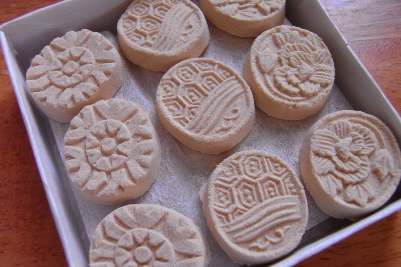 A box lined with white paper holding nine round, pale beige dry sweets, each pressed with a pattern such as a flower, a tortoiseshell hexagon design or waves