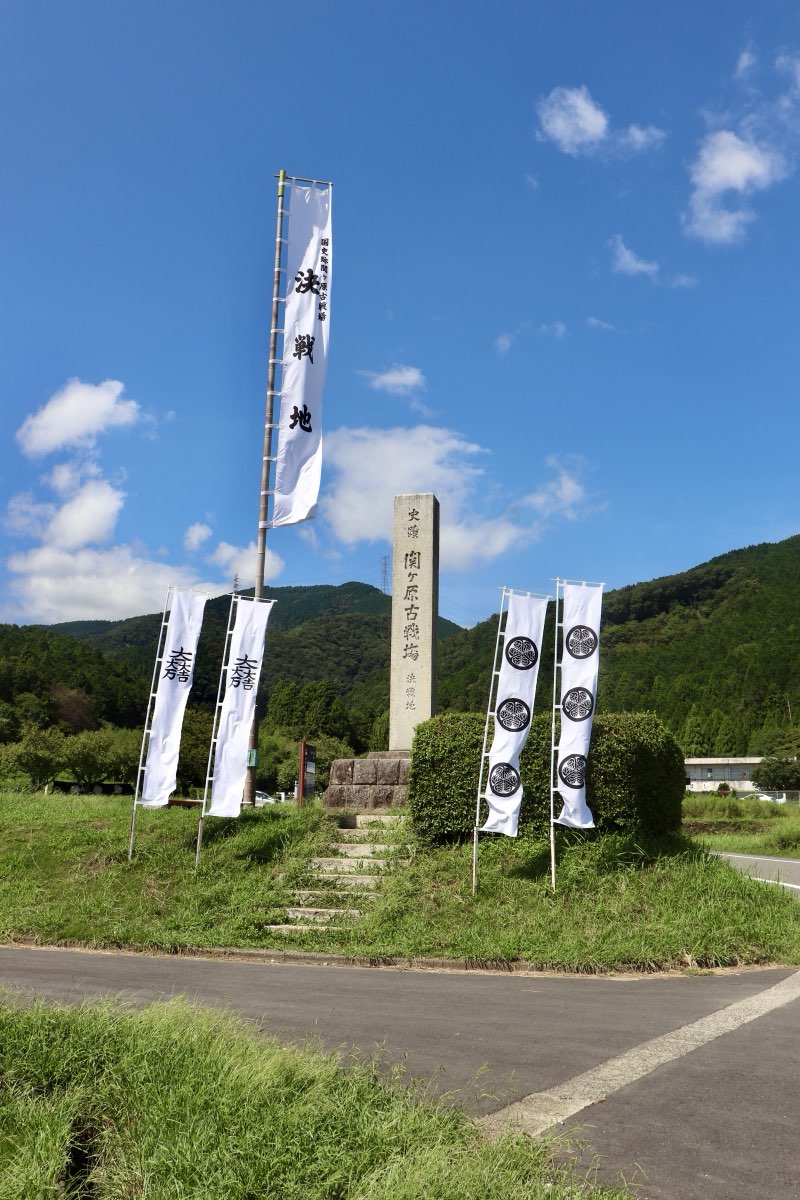 A tall stone marker inscribed as the Sekigahara battlefield decisive battle site, on a grassy mound with stone steps, flanked by white banners, with green hills and a blue sky behind