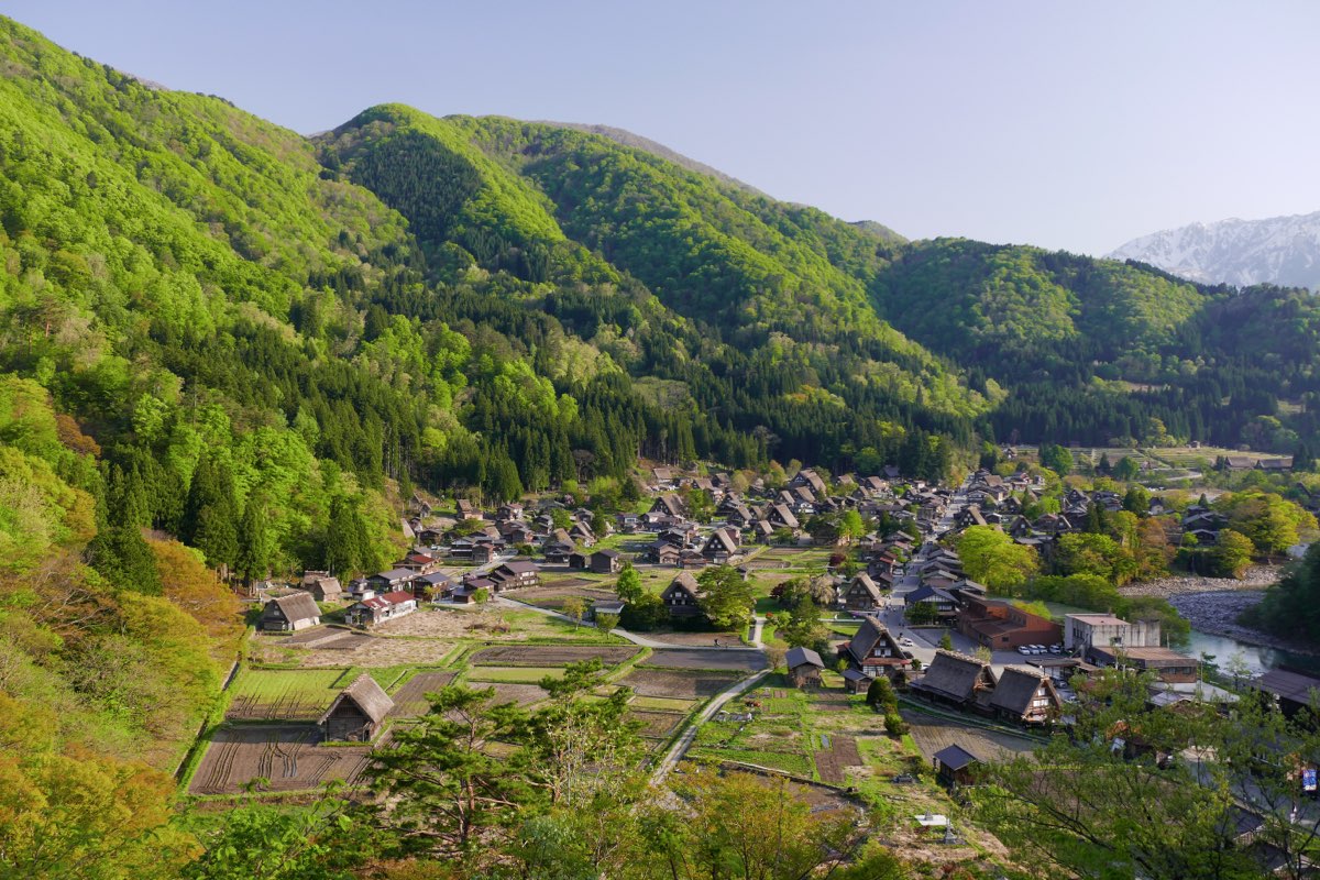 A valley village of steep thatched-roof houses among fields and trees, seen from above, with green forested mountains behind and a snow-capped peak in the distance