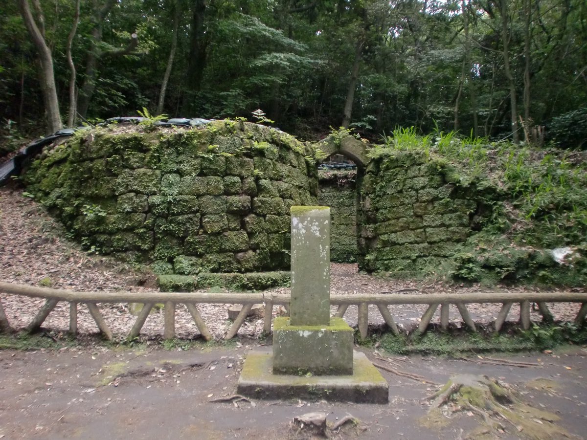 A low, rounded structure of large moss-covered stone blocks in a wood, with a narrow opening under an arched stone on the right, seen across a low wooden fence; a weathered square stone post on a stepped base stands in front on a dirt path, and tall trees fill the background