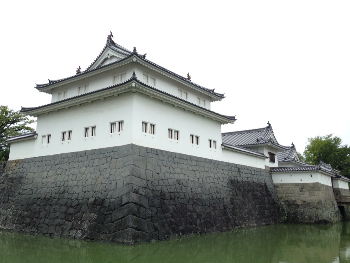 A white corner turret with two tiers of grey tiled roofs standing on a high dark stone wall that rises straight from a green moat, with lower white gate buildings with tiled roofs continuing to the right on lower stone walls, under an overcast sky