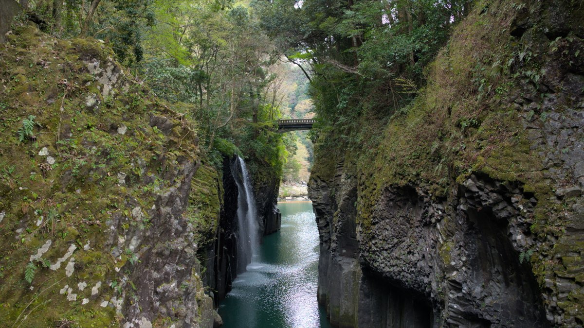 A narrow river gorge with dark, moss-covered rock walls of vertical columns; a thin waterfall drops from the left cliff into blue-green water, and an arched bridge crosses the gorge in the distance among trees