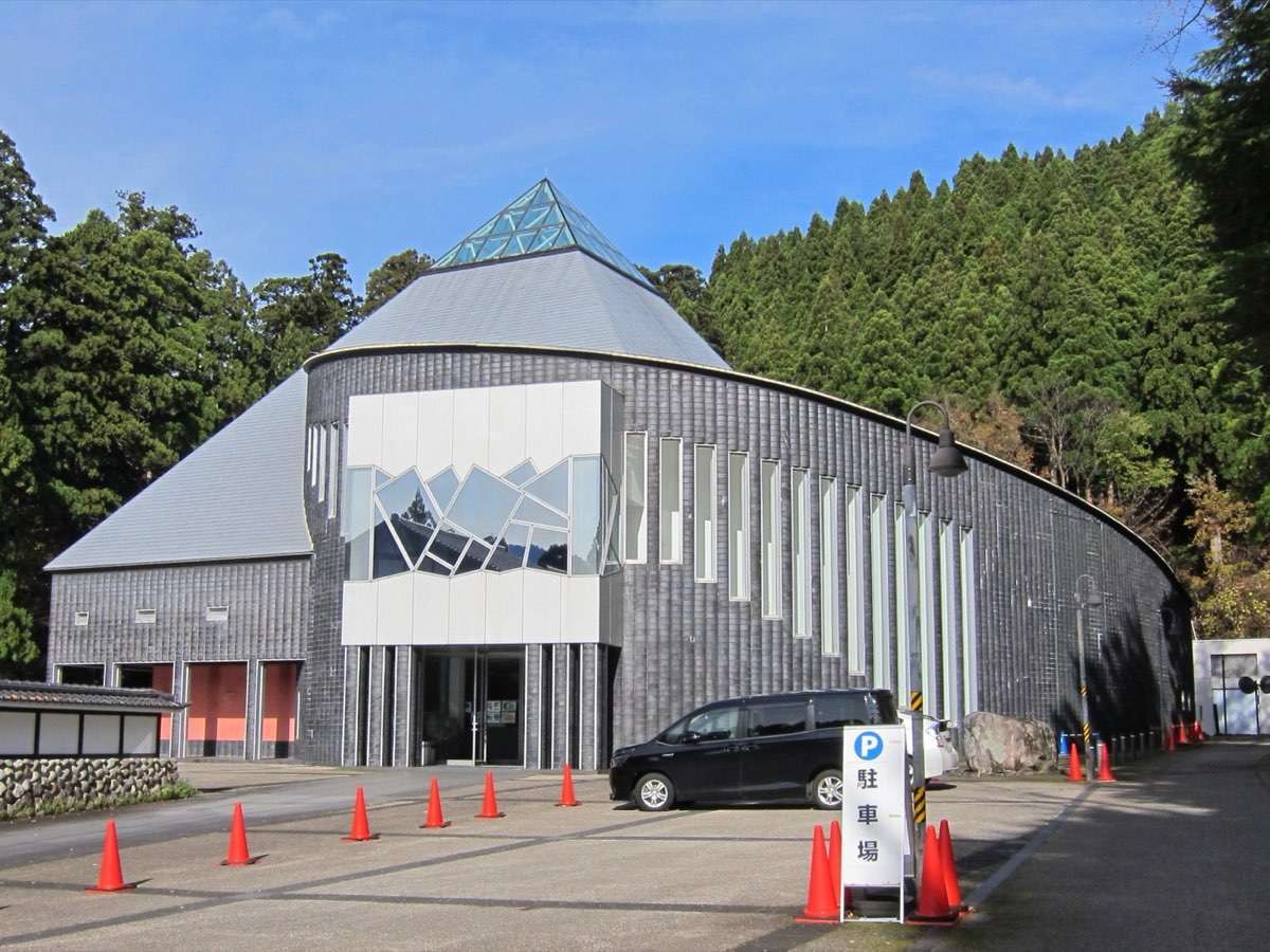 A modern museum building with a curved grey wall, a pyramid-shaped roof and a glass skylight, in front of a forested hillside, photographed in 2018