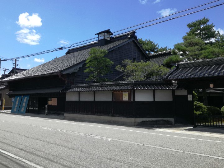 The dark wooden two-storey main house of Kanaoka-tei with a tiled roof and a long wall with a tiled roof along a city street in Toyama, photographed in 2018