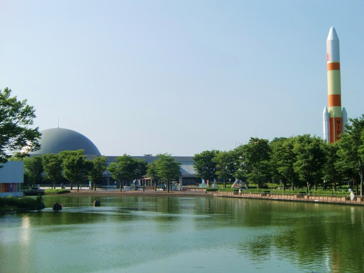 A pond in a park with trees along its edge; across the water stand the low Tsukuba Expo Center building with a large grey planetarium dome and, on the right, a tall orange and white rocket model against a clear sky