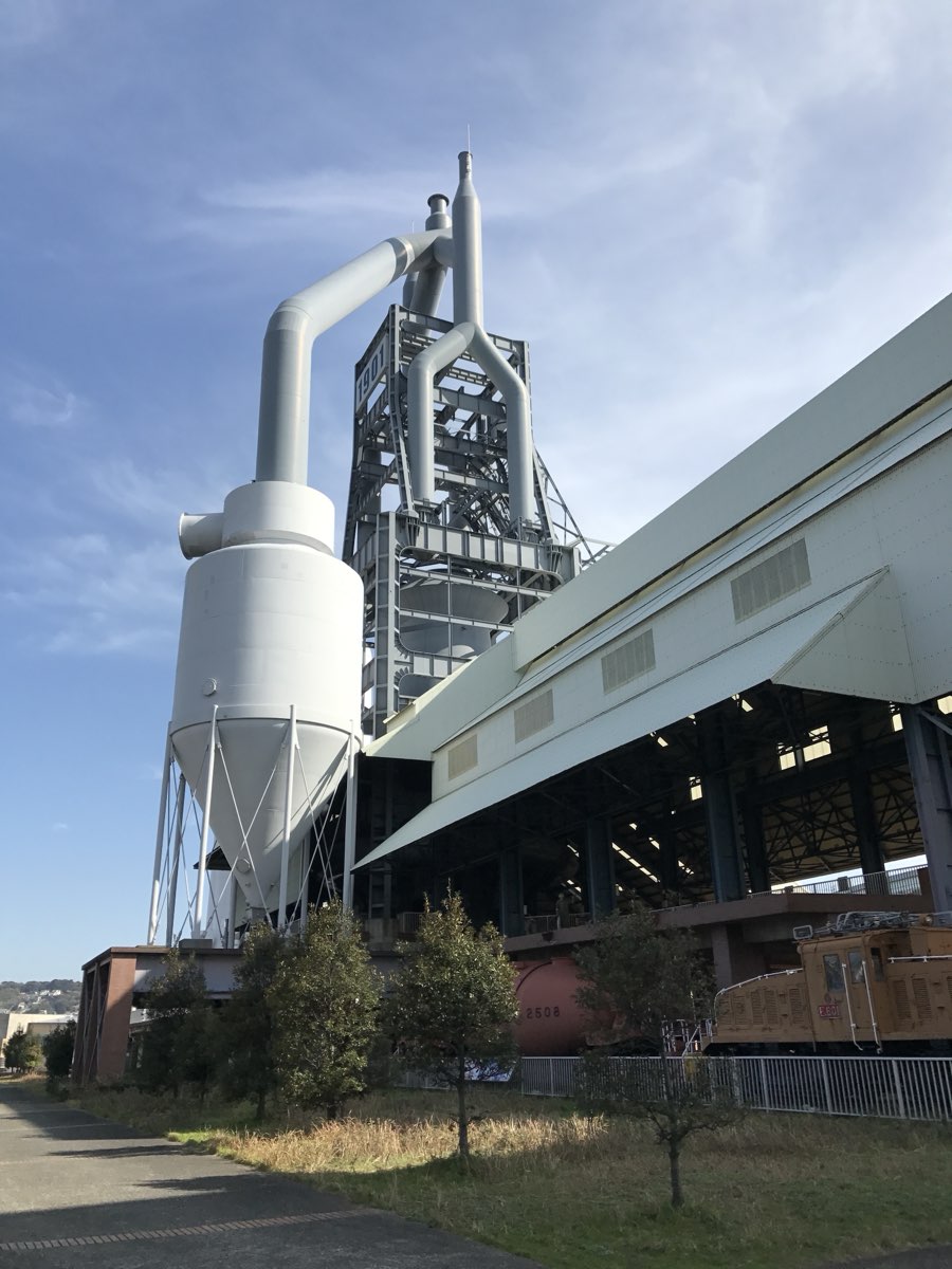 A tall steel blast furnace tower with large grey pipes and a cone-shaped dust collector, beside a long pale building with an open ground floor and an old electric locomotive, seen from a path under a blue sky
