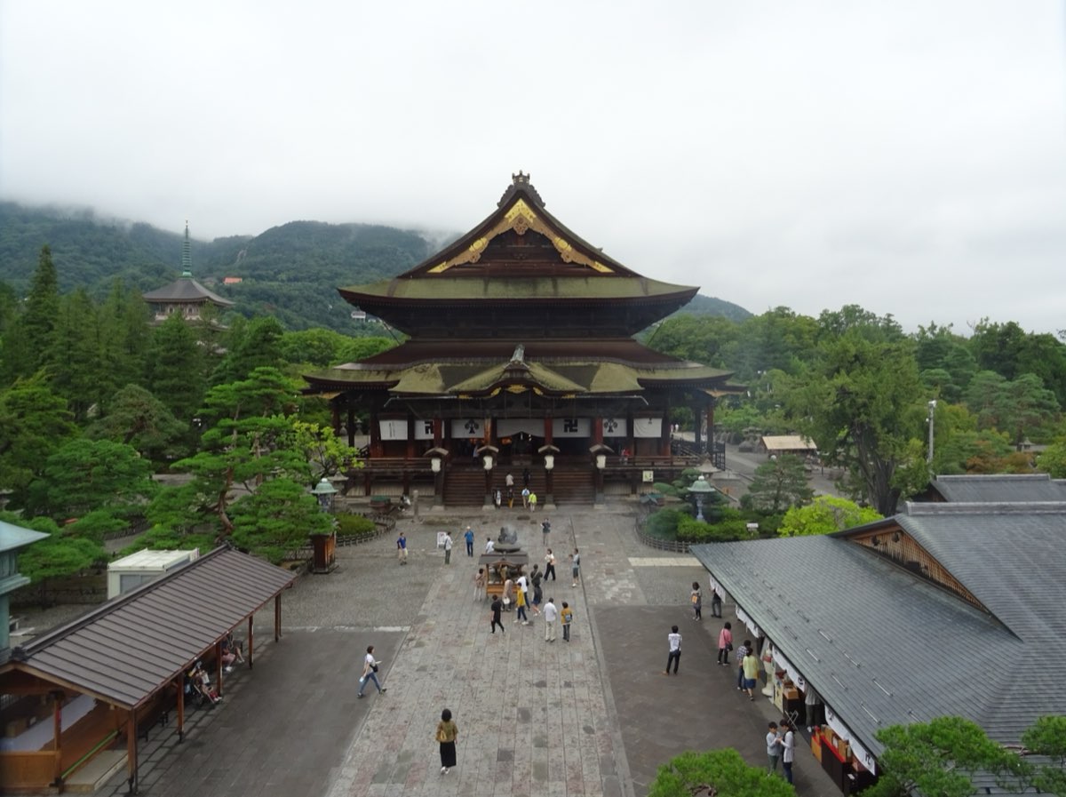 The large two-roofed wooden main hall of Zenkōji seen from above, with a paved precinct in front, pine trees around it and wooded hills behind, photographed in September 2018